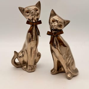 May include: Two silver-toned metallic cat figurines in a seated position. Each cat features pointed ears, amber-colored eyes, and a dark brown ribbon tied in a bow around its neck. The figures have a smooth, reflective surface and upright posture.