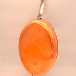 May include: An oval-shaped copper pan with a polished metallic finish. The pan features a long, straight silver-toned metal handle attached at the top and a smaller, curved silver-toned metal loop handle positioned at the bottom edge.