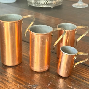 May include: A set of four graduated copper measuring cups with cylindrical bodies and thin, curved brass-toned handles. Each cup features a polished copper exterior and a metallic interior lining, arranged in descending order of size from left to right.