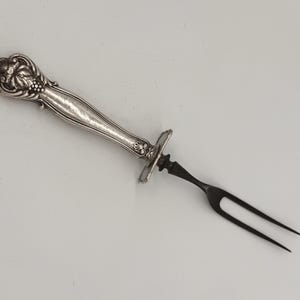 May include: A silver-toned metal serving fork featuring an ornate, embossed handle with floral and fruit motifs. The handle transitions into a smooth, slender neck, ending in a two-pronged, dark-colored metal fork head with sharp, pointed tips.
