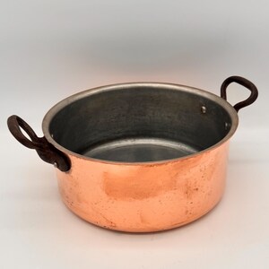 May include: A round copper pot with a metallic silver-toned interior lining. The exterior features a polished copper finish with visible surface variations. Two dark, textured metal handles are attached to opposite sides of the pot rim.
