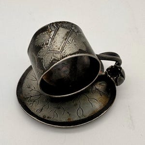 May include: A tarnished silver-toned metal cup and saucer set. The cup features an engraved rectangular plaque surrounded by leaf motifs. The saucer displays a delicate etched branch pattern. A small decorative flower-shaped detail adorns the handle.