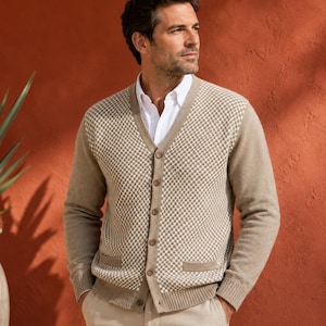 Puede incluir: Un cárdigan beige y blanco con cuello en V, frente de punto con patrón de diamantes, mangas largas, puños y dobladillo acanalados y cierre frontal de botones. La prenda se lleva sobre una camisa blanca de cuello y pantalones beige.