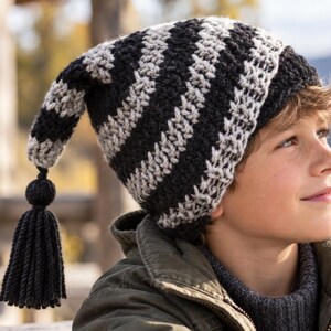 May include: A slouchy knit beanie featuring alternating horizontal stripes of black and light gray yarn. The hat has a long, pointed tail that hangs down, finished with a solid black yarn pom-pom and a long, thick black yarn tassel at the end.