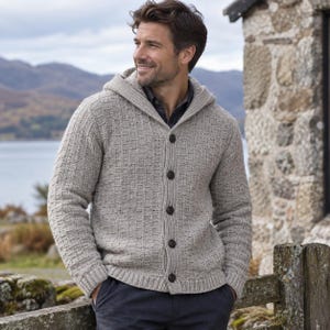 Puede incluir: Un cárdigan beige con capucha, patrón de punto texturizado, cierre frontal con cinco botones de color marrón oscuro, mangas largas y dobladillo acanalado. La prenda se lleva sobre una camisa de cuello oscuro y pantalones oscuros.