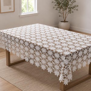Puede incluir: Un mantel rectangular blanco que presenta un intrincado patrón de encaje de ganchillo repetitivo con motivos florales. La tela tiene un diseño calado delicado con bordes festoneados que cuelgan sobre los lados de una mesa de madera.