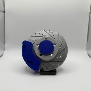 May include: A silver-colored circular brake rotor featuring a series of drilled holes and diagonal ventilation slots. A solid blue brake caliper is attached to the side of the rotor, and a circular blue hub cap is positioned in the center of the disc.