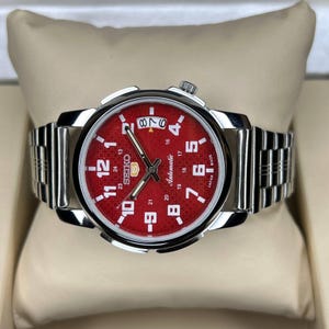 May include: A silver-tone stainless steel Seiko 5 automatic wristwatch featuring a vibrant red dial with a textured pattern, white Arabic numerals, a date window at the three o'clock position, and a metallic link bracelet with a polished finish.
