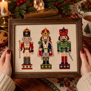 Könnte beinhalten: Ein gerahmtes Kreuzstichbild mit drei Nussknacker-Figuren auf cremefarbenem Stoff. Die linke Figur trägt eine schwarze Mütze und eine rote Uniform, die mittlere Figur eine Krone und einen blauen Umhang, und die rechte Figur eine rote Mütze.