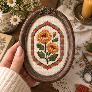 以下が含まれることがあります： 緑の葉を持つ2つの黄色い花がクロスステッチで刺繍された楕円形の木製フレーム。刺繍はクリーム色の布地の上に赤と緑の四角いチェック模様で縁取られています。フレームはダークカラーの仕上げが施されています。