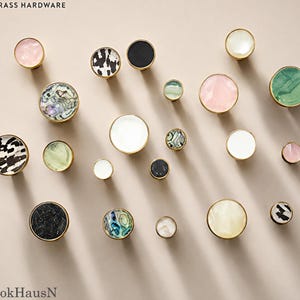 May include: A collection of round cabinet knobs with brass hardware edges, featuring various inlaid surfaces including pink quartz, black and white patterns, iridescent abalone shell, solid white, solid black, and green marbled stone textures.
