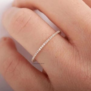 Könnte beinhalten: Ein dünner Ring aus Roségold mit einer durchgehenden Reihe kleiner, klarer, rund geschliffener Steine in Pavé-Fassung auf der Oberseite. Das Metall hat ein warmes, metallisches Finish und die Steine sind in einer geraden, gleichmäßigen Linie angeordnet.
