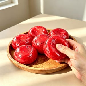 May include: Six round, glossy, bright red objects with a smooth, slightly translucent finish and dark speckles, arranged on a circular wooden tray. Each object has a small, shallow indentation in the center, resembling a small, firm, round fruit.