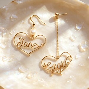 Op de afbeelding: Een paar goudkleurige draadorbellen met hartvormige frames. De ene oorbel toont de naam Jane met een kleine witte parel aan een haakje, terwijl de andere de naam Gigi heeft hangend aan een lange gouden ketting met een stekersluiting.