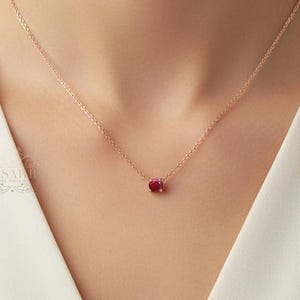May include: A delicate rose gold chain necklace featuring a single, round, faceted deep red gemstone pendant. The necklace rests against the collarbone, showcasing a minimalist design with a thin metal chain and a secure prong setting for the stone.