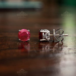 Könnte beinhalten: Ein Paar Ohrstecker mit runden, facettierten, tiefroten Edelsteinen in silberfarbenem Metall. Ein Ohrring ist von vorne zu sehen und zeigt den runden Stein, während der andere von der Seite abgebildet ist und die filigrane Metallarbeit offenbart.