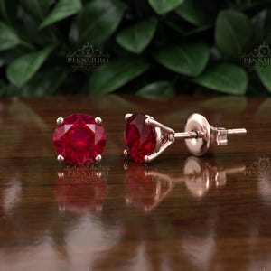 Könnte beinhalten: Ein Paar Ohrstecker mit rund geschliffenen, leuchtend roten Edelsteinen in einer Fassung aus Roségold. Ein Ohrring ist von vorne zu sehen, der andere von der Seite, wobei die vierzackige Fassung und der Stiftverschluss deutlich erkennbar sind.