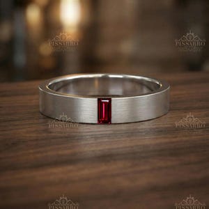 May include: A brushed silver-tone band ring featuring a single rectangular baguette-cut red gemstone set vertically in the center. The ring has a flat profile and a smooth, matte metallic finish, with the brand name PISSARRO visible in the image.