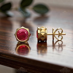 Könnte beinhalten: Ein Paar goldene Ohrstecker mit einem runden, facettierten, tiefrosa Edelstein in einer sechseckigen Fassung. Die Seite des Ohrrings zeigt ein pyramidenförmiges Muster, und der Verschluss ist als dekorativer, durchbrochener Gold-Schmetterling gestaltet.