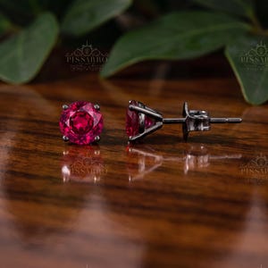 Puede incluir: Un par de pendientes de botón con gemas rojas redondas y facetadas, engastadas en garras de metal de tono gris oscuro. Un pendiente se muestra de frente, mientras que el otro está posicionado para mostrar el perfil lateral y el cierre.