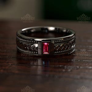 May include: A dark metallic band ring featuring a central rectangular baguette-cut pink gemstone. The band displays a textured, woven-style pattern bordered by rows of small, raised metallic beads. The text PISSARRO is visible on the ring.