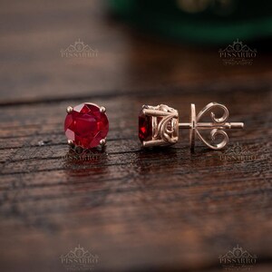 May include: A pair of stud earrings featuring round, faceted deep red gemstones set in rose gold. One earring is shown from the front, while the other is viewed from the side, revealing an intricate, scroll-like metal gallery and a secure post back.