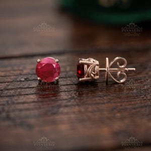 Könnte beinhalten: Ein Paar Ohrstecker mit runden, facettierten, tiefroten Edelsteinen in einer Fassung aus Roségold. Ein Ohrring ist von vorne zu sehen und zeigt den runden Stein, während der andere von der Seite abgebildet ist, was die filigrane Metallfassung mit Schnörkeln und den Stift offenbart.