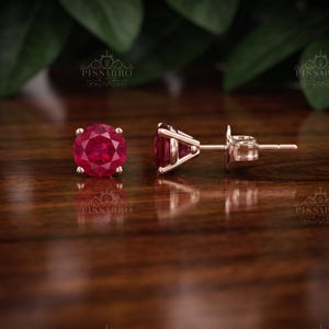 Könnte beinhalten: Ein Paar Ohrstecker mit rund geschliffenen, tiefroten Rubinen in Roségold gefasst. Ein Ohrring ist von vorne zu sehen und zeigt den facettierten Edelstein, während der andere so positioniert ist, dass das Seitenprofil und der Metallstift sichtbar sind.