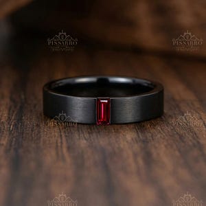 Könnte beinhalten: Ein mattschwarzer Ring mit einem einzelnen, rechteckigen, im Baguetteschliff geschliffenen roten Edelstein, der horizontal in der Mitte gefasst ist. Die Ringschiene hat eine glatte, gebürstete Oberfläche und ein flaches Profil, mit dem PISSARRO-Logo im Hintergrund.