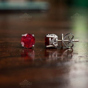 May include: A pair of stud earrings featuring round, faceted, deep red gemstones set in silver-toned metal. One earring is shown from the front, while the other is viewed from the side, revealing an intricate, scroll-like metalwork design on the setting.