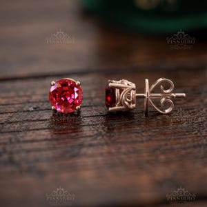 Könnte beinhalten: Ein Paar Ohrstecker mit runden, leuchtend roten Edelsteinen in Roségold gefasst. Ein Ohrring ist von vorne zu sehen und zeigt den facettierten roten Stein, während der andere von der Seite abgebildet ist und das filigrane Metallmuster zeigt.