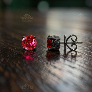 Pode incluir: Um par de brincos de tarracha com pedras vermelhas redondas e facetadas em metal escuro. Um brinco é visto de frente, enquanto o outro revela o perfil lateral e o intrincado trabalho de volutas da parte de trás do metal.