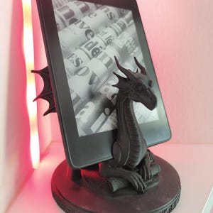 Könnte beinhalten: Ein schwarzer E-Reader, der von einem mattschwarzen Drachenständer gehalten wird. Der Drache sitzt auf einem runden Sockel mit eingravierten Mustern. Der Bildschirm des E-Readers zeigt ein Graustufenbild mit überlappendem Zeitungstext.