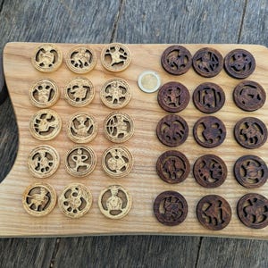 Könnte beinhalten: Ein Set aus dreißig runden Spielsteinen aus Holz mit filigranen, ausgeschnittenen Sternzeichen, angeordnet in zwei Gruppen zu je fünfzehn auf einem hellen Holzbrett. Eine Gruppe ist aus hellem Holz, die andere aus dunklem Holz, mit einer Münze.