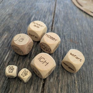 Puede incluir: Una colección de dados de madera de color claro con palabras en alemán grabadas como Schokolade, Apfel, Hobby, Disco, Kneipe, ja, jain, ICH, DU, sofort y morgen, junto a dos dados más pequeños que muestran gestos de mano con el signo de paz.