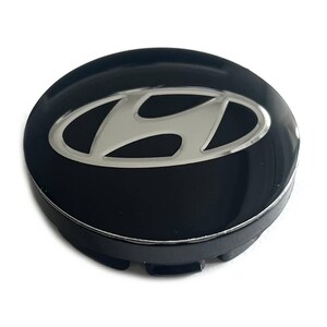May include: A circular black wheel center cap featuring a silver Hyundai logo in the center. The cap has a glossy finish and a raised edge, with visible mounting clips on the underside. The design is simple, round, and metallic in appearance.