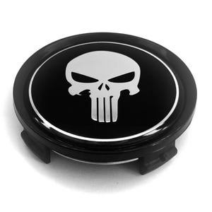 May include: A circular black wheel center cap featuring a silver-toned skull emblem in the center. The emblem is surrounded by a thin silver circular border. The cap has a glossy finish and includes mounting clips on the underside.