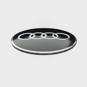 May include: A circular black emblem featuring the silver Audi logo, which consists of four interlocking rings. The emblem has a smooth, glossy finish with a silver border around the outer edge, presented from a slightly elevated perspective.