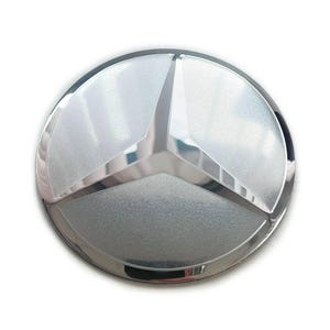 May include: A circular silver-toned emblem featuring a three-pointed star design enclosed within a ring. The surface of the emblem displays a metallic, reflective finish with a subtle textured grain across the three triangular segments.