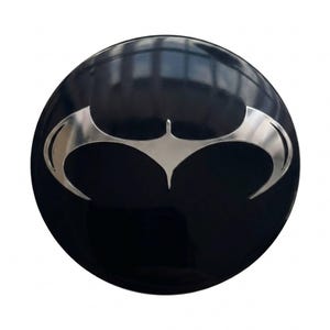 Op de afbeelding: Een rond embleem met een glanzend zwarte afwerking en een centraal geplaatst, metallic zilveren vleermuislogo. Het ontwerp bestaat uit een gestileerd, symmetrisch vleermuissilhouet met puntige vleugels en een centrale piek op een glad oppervlak.