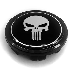 May include: A circular black wheel center cap featuring a silver-colored skull emblem in the center. The emblem is surrounded by a thin silver ring, and the cap has a glossy finish with a raised edge and mounting clips visible on the underside.