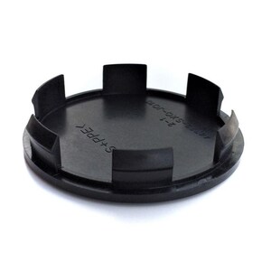 May include: A circular black plastic wheel center cap featuring six evenly spaced mounting clips along the outer edge. The interior surface is embossed with the text S+PPE and 44732-SXO-J010 2-1, indicating specific part identification details.