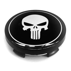 May include: A circular black wheel center cap featuring a white stylized skull graphic in the center. The cap has a glossy finish with a thin white circular border surrounding the skull design and includes black mounting clips on the underside.