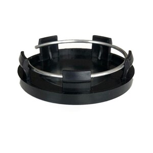 May include: A circular black plastic wheel center cap featuring a smooth, flat top surface and a series of vertical mounting clips along the perimeter. A thin, silver-colored metal tension ring is fitted around the clips to secure the cap.
