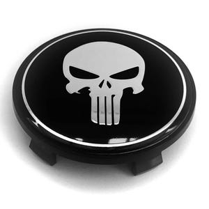 May include: A circular black wheel center cap featuring a silver-colored skull emblem in the center. The emblem has a stylized design with sharp eye sockets and vertical teeth. A thin silver ring outlines the edge of the glossy black surface.