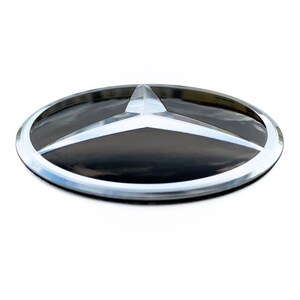 May include: A circular Mercedes-Benz emblem featuring a glossy black background enclosed by a polished silver-toned metallic ring. The iconic three-pointed star design is rendered in a raised, reflective silver finish, centered within the circular frame.