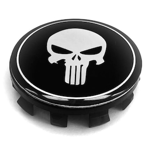 Könnte beinhalten: Eine runde schwarze Radnabenkappe mit einer weißen, stilisierten Totenkopfgrafik in der Mitte. Die Kappe hat eine glänzende Oberfläche mit einem dünnen weißen kreisförmigen Rand um das Totenkopfdesign und schwarzen Kunststoff-Befestigungsclips.