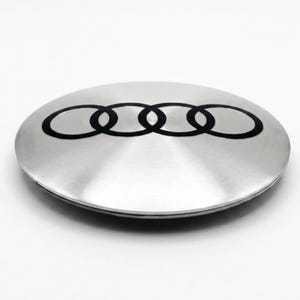 May include: A circular, silver-toned metal wheel center cap featuring the black Audi four-ring logo centered on its brushed surface. The object has a smooth, slightly domed profile and a polished metallic finish, displaying the iconic automotive brand emblem clearly across the top.