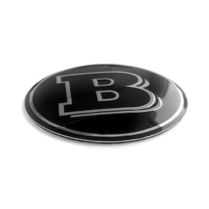 May include: A circular black emblem featuring a stylized silver letter B in the center. The design includes a thin silver border around the edge and a reflective, glossy finish on the surface, creating a smooth and polished appearance.