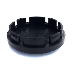 May include: A circular black plastic wheel center cap featuring a series of evenly spaced mounting clips along the outer edge. The interior surface is embossed with the letters JD, the code JA601, and the characters 15A.
